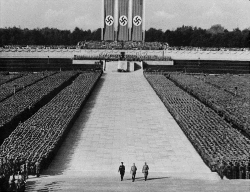 Still of Hitler and two other figures walking through middle of massed ranks of soliders. Nazi flags in background.